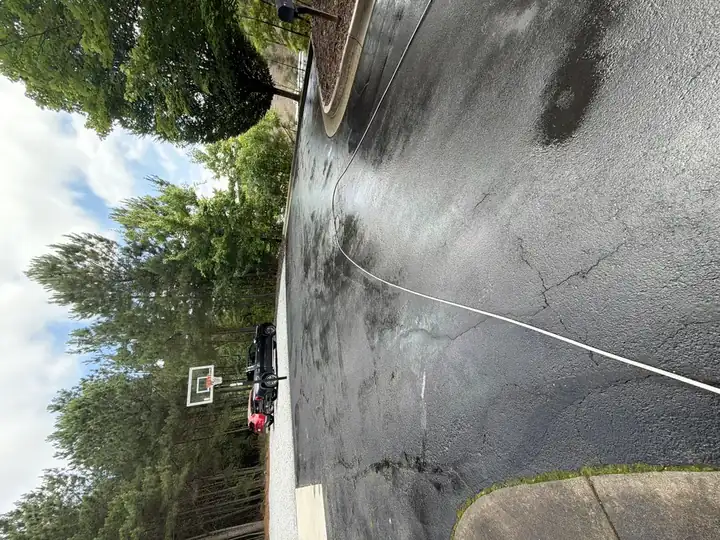 Wet asphalt driveway and basketball court area during pressure washing in Alpharetta, GA, with hose visible and trucks parked in the background