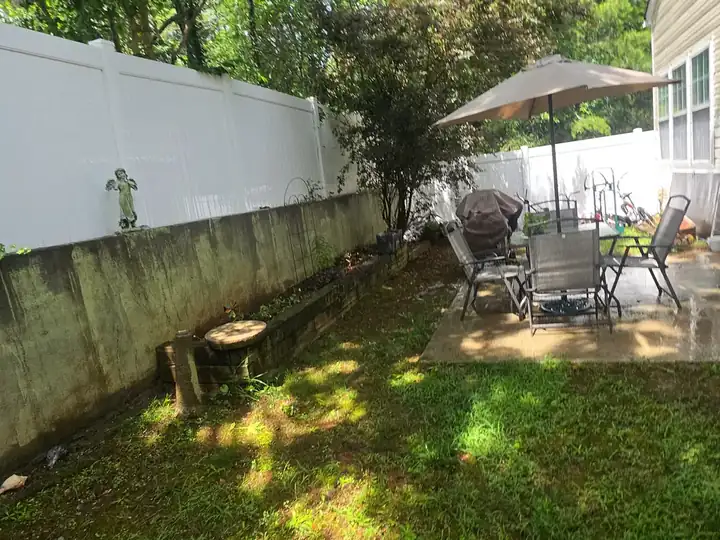 Backyard with a concrete patio, outdoor furniture, and a concrete retaining wall with algae growth below a vinyl fence at a pressure washing job in Woodstock, GA.