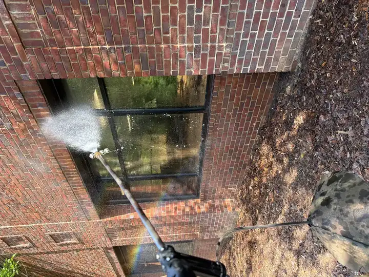 Extension wand spraying water on commercial brick building windows during pressure washing service in Alpharetta, GA, with rainbow visible in the mist